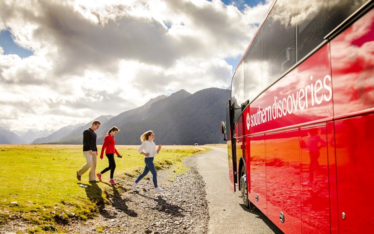 Tourists boarding a red coach to Milford Sound, New Zealand, with mountains in the background.
