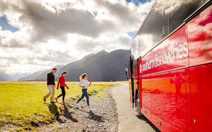 Tourists boarding a red coach to Milford Sound, New Zealand, with mountains in the background.