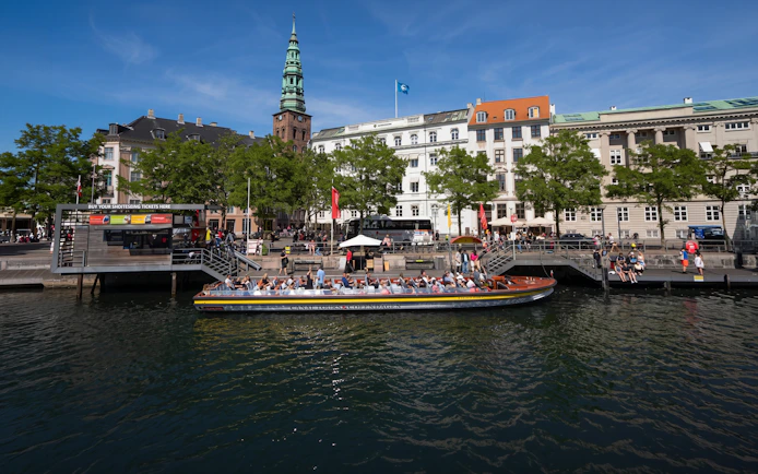 Sightseeing boat in Copenhagen canal with historic buildings and spire in background.