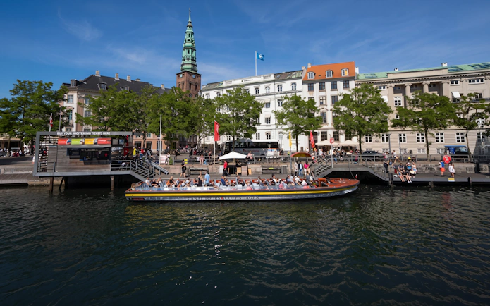 Sightseeing boat in Copenhagen canal with historic buildings and spire in background.