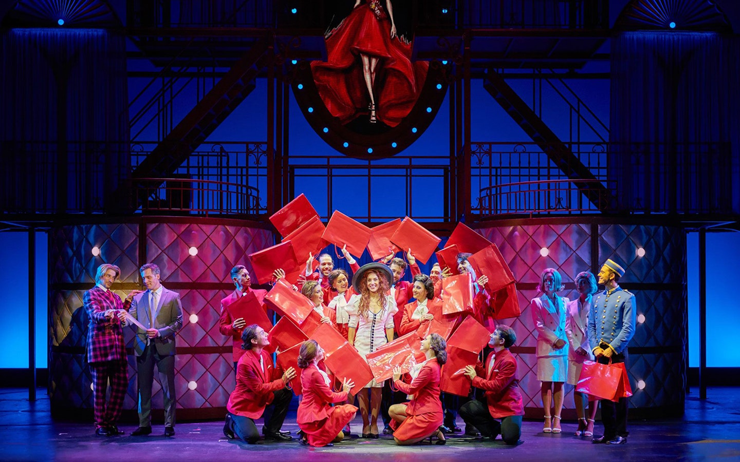 Cast performing a scene from "Pretty Woman - El musical" with red shopping bags on stage.