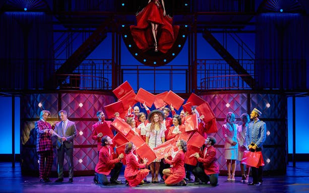 Cast performing a scene from "Pretty Woman - El musical" with red shopping bags on stage.