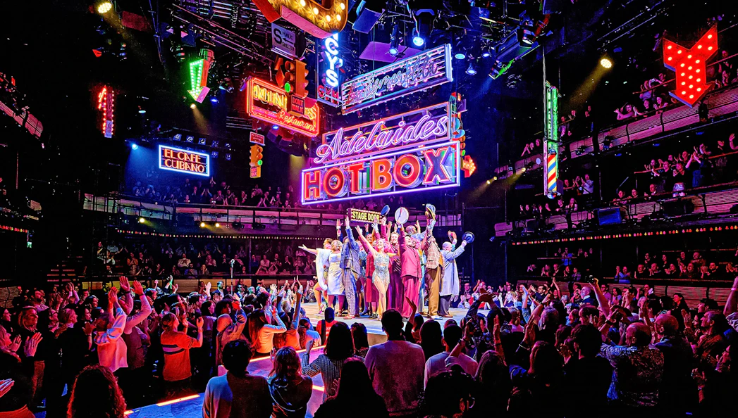 Cast of "Guys & Dolls" performing on stage with vibrant neon signs and audience in London theater.