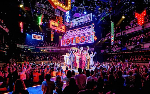 Cast of "Guys & Dolls" performing on stage with vibrant neon signs and audience in London theater.