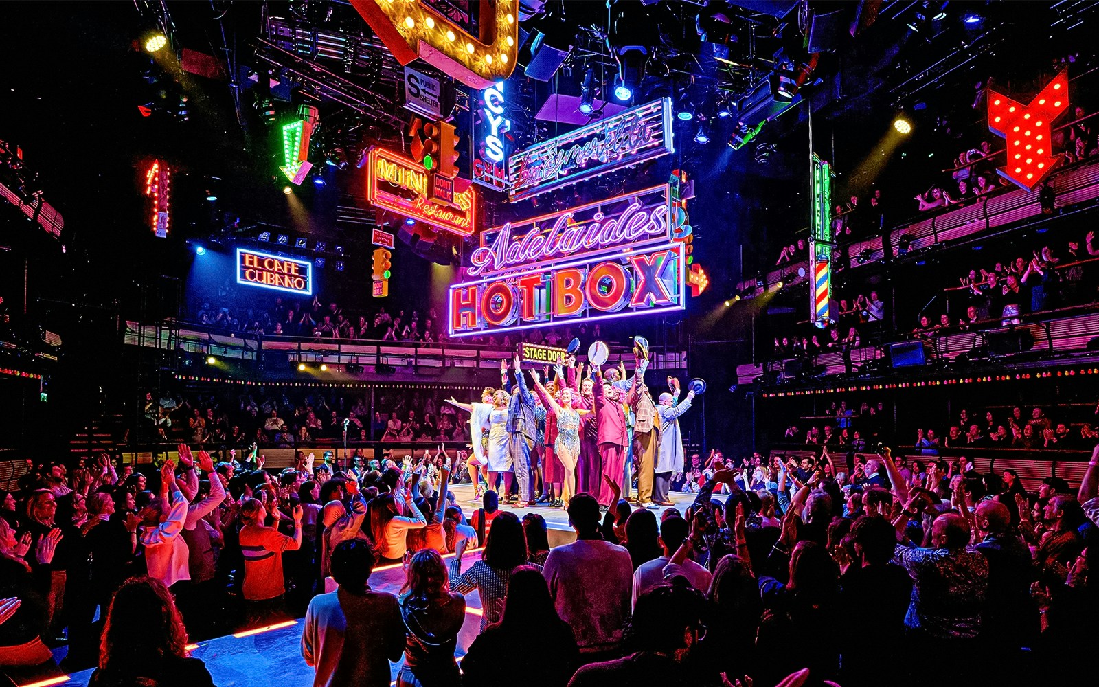 Cast of "Guys & Dolls" performing on stage with vibrant neon signs and audience in London theater.