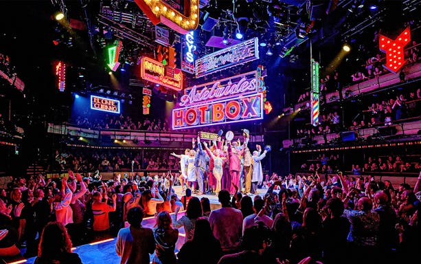 Cast of "Guys & Dolls" performing on stage with vibrant neon signs and audience in London theater.