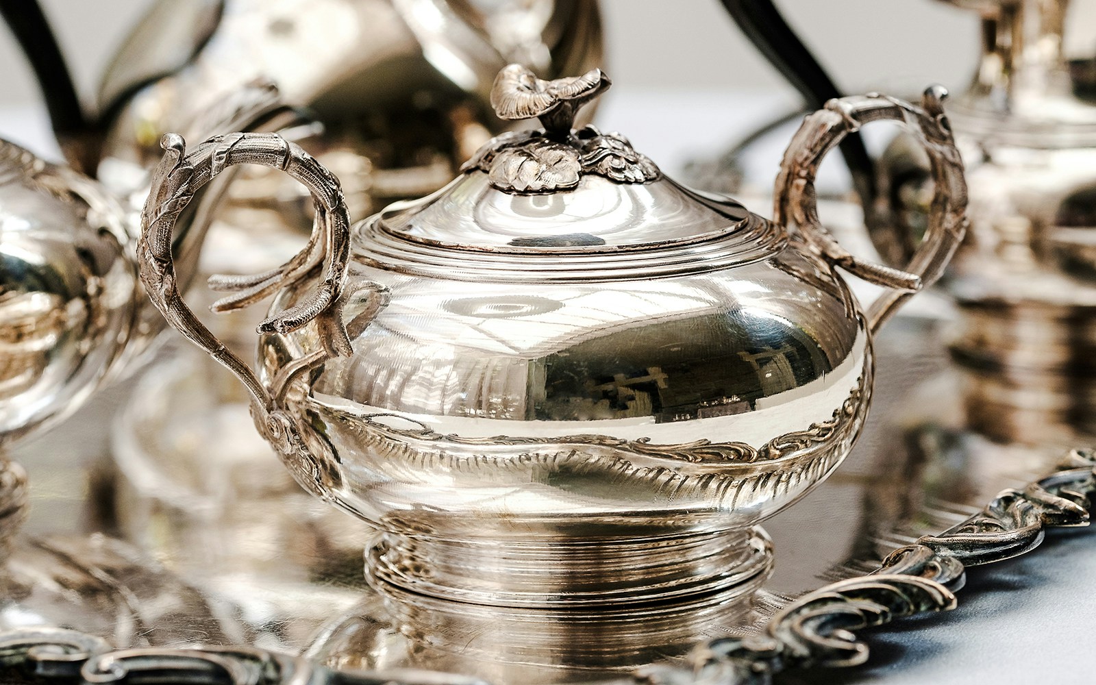 Antique silver teapot with ornate handles and lid on a reflective tray.