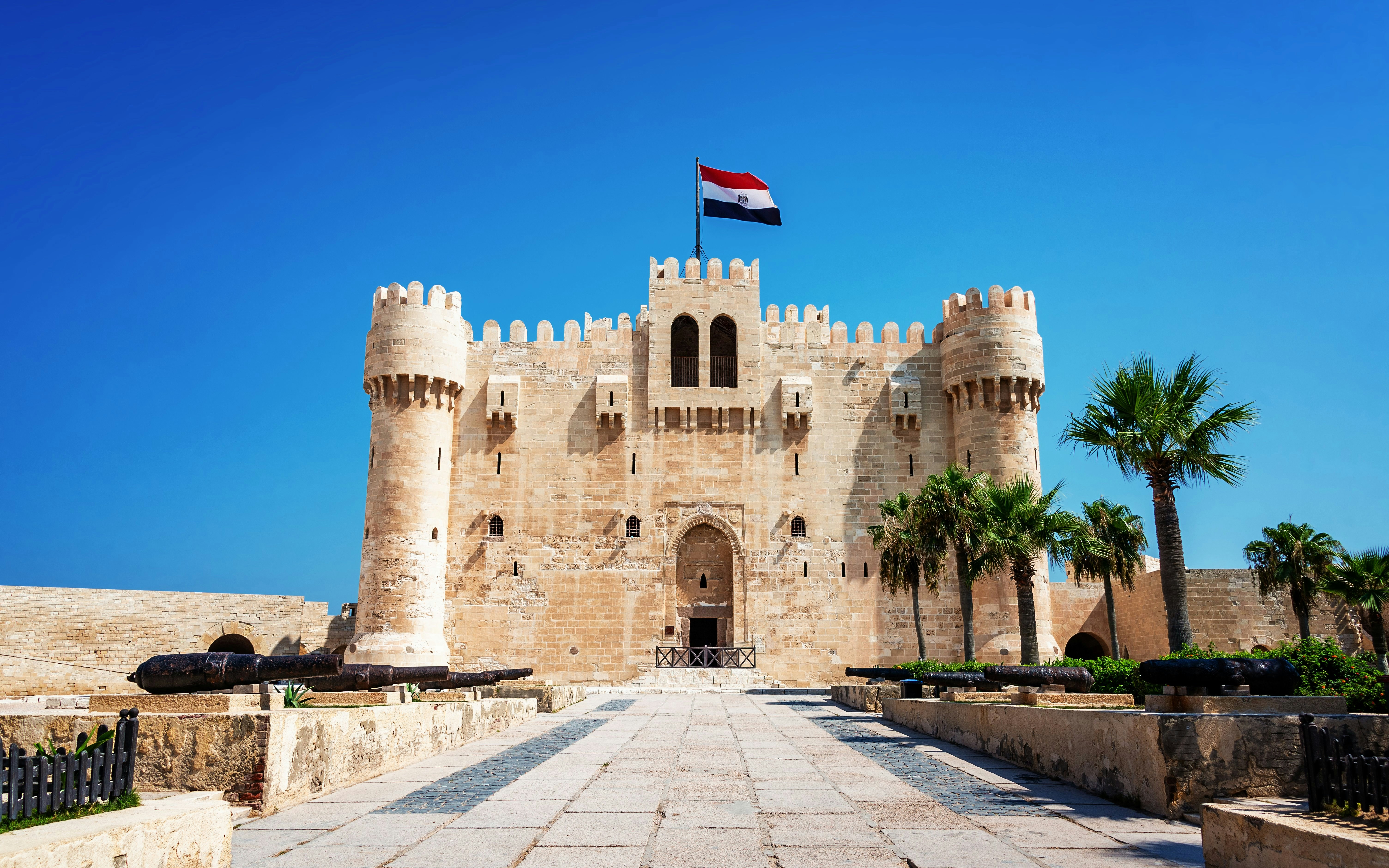 Citadel of Qaitbay in Alexandria, Egypt, with Egyptian flag and palm trees.
