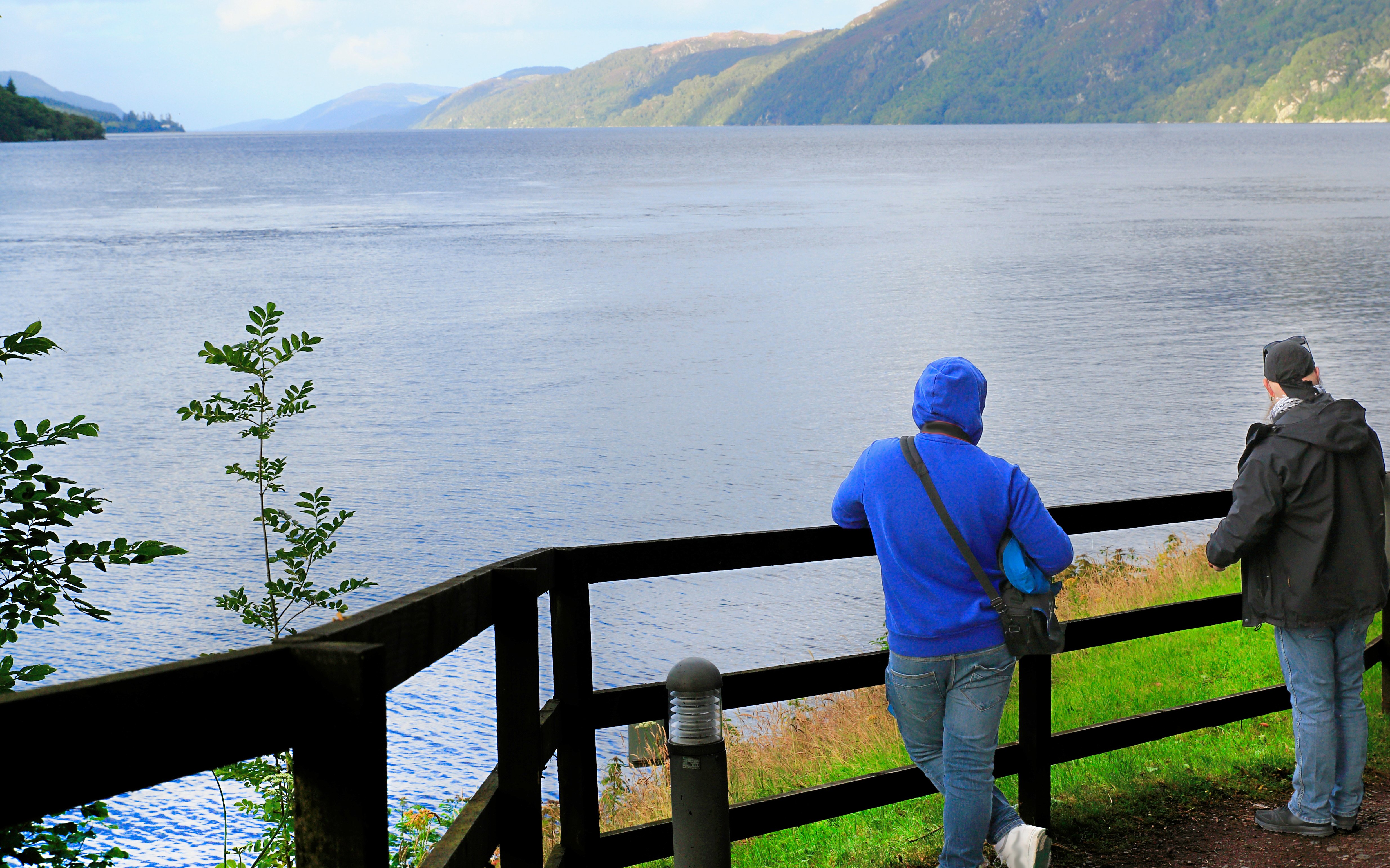 Tourists viewing Loch Ness from a wooden fence viewpoint in Scotland.