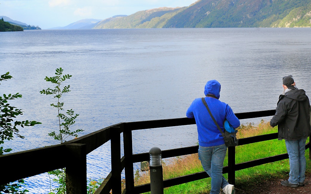 Tourists viewing Loch Ness from a wooden fence viewpoint in Scotland.