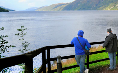 Tourists viewing Loch Ness from a wooden fence viewpoint in Scotland.