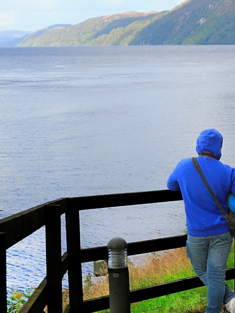 Tourists viewing Loch Ness from a wooden fence viewpoint in Scotland.