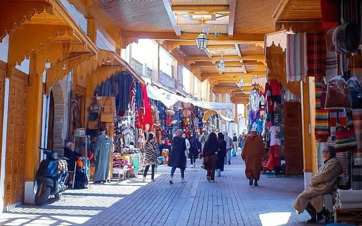 Bustling Marrakech market with people shopping, colorful textiles, and traditional architecture.