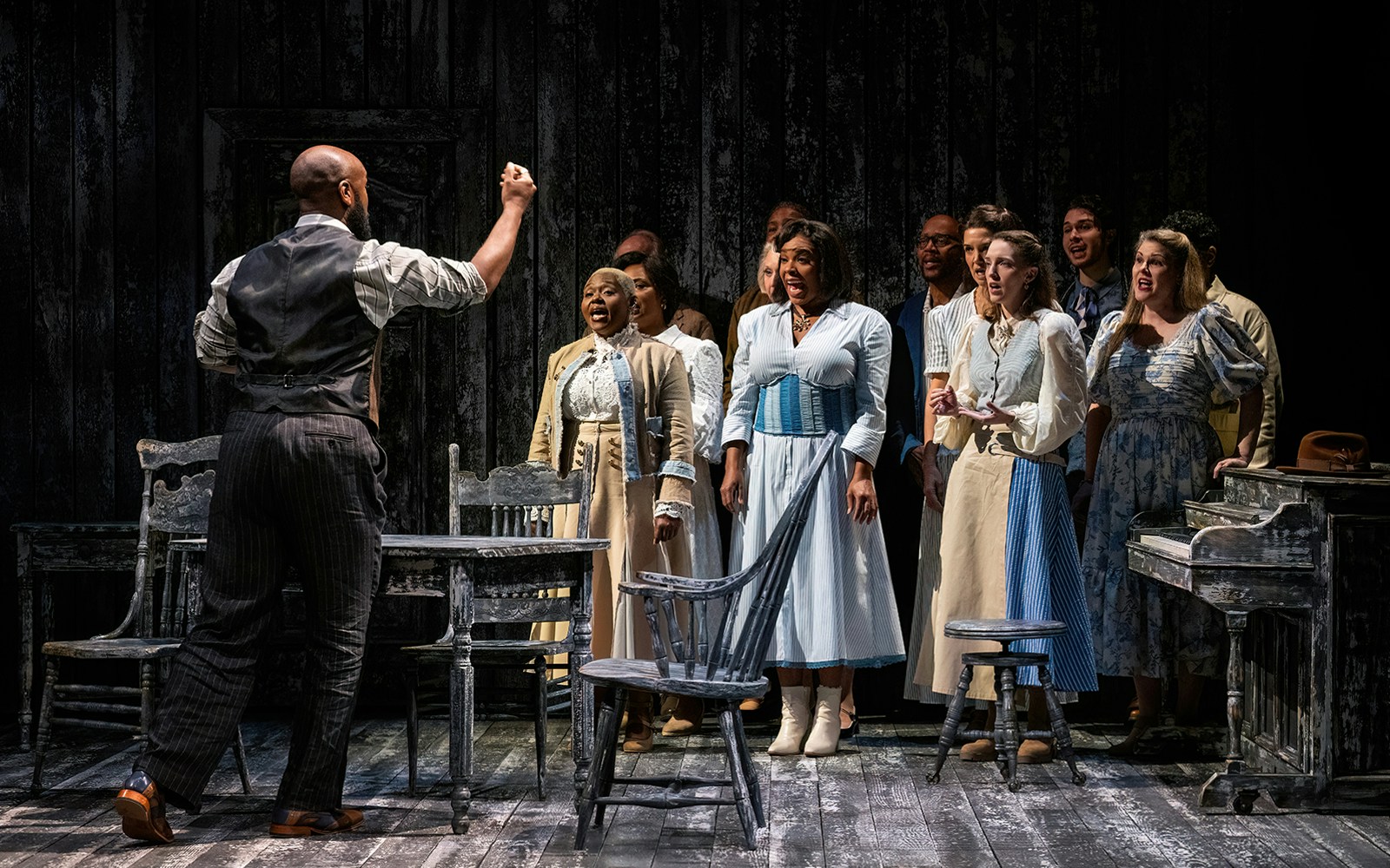 Cast performing a scene from "Our Town" on stage with a wooden table and chairs.