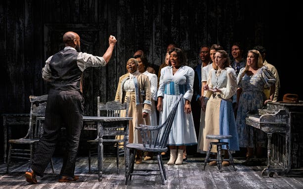 Cast performing a scene from "Our Town" on stage with a wooden table and chairs.