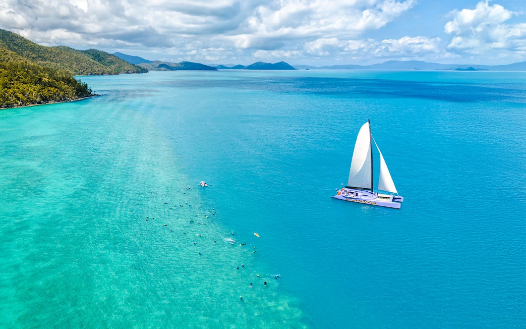 Sailing catamaran on turquoise waters near Airlie Beach with snorkelers in the ocean.