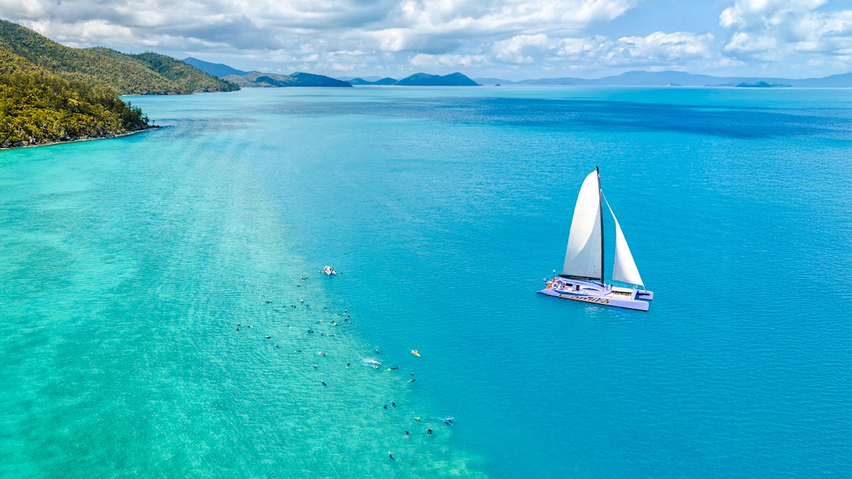 Sailing catamaran on turquoise waters near Airlie Beach with snorkelers in the ocean.