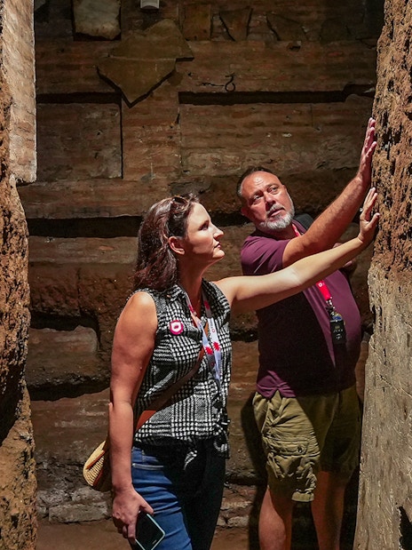 Tourists exploring narrow passage in Roman Catacombs, touching ancient walls.