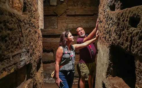 Tourists exploring narrow passage in Roman Catacombs, touching ancient walls.