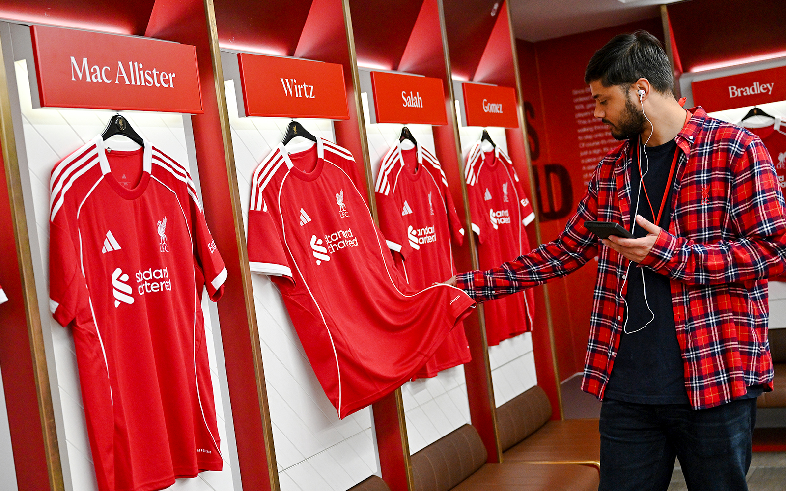 Liverpool FC jerseys in Anfield dressing room with visitor examining a shirt.