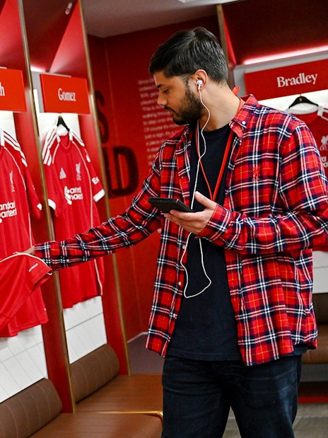 Liverpool FC jerseys in Anfield dressing room with visitor examining a shirt.