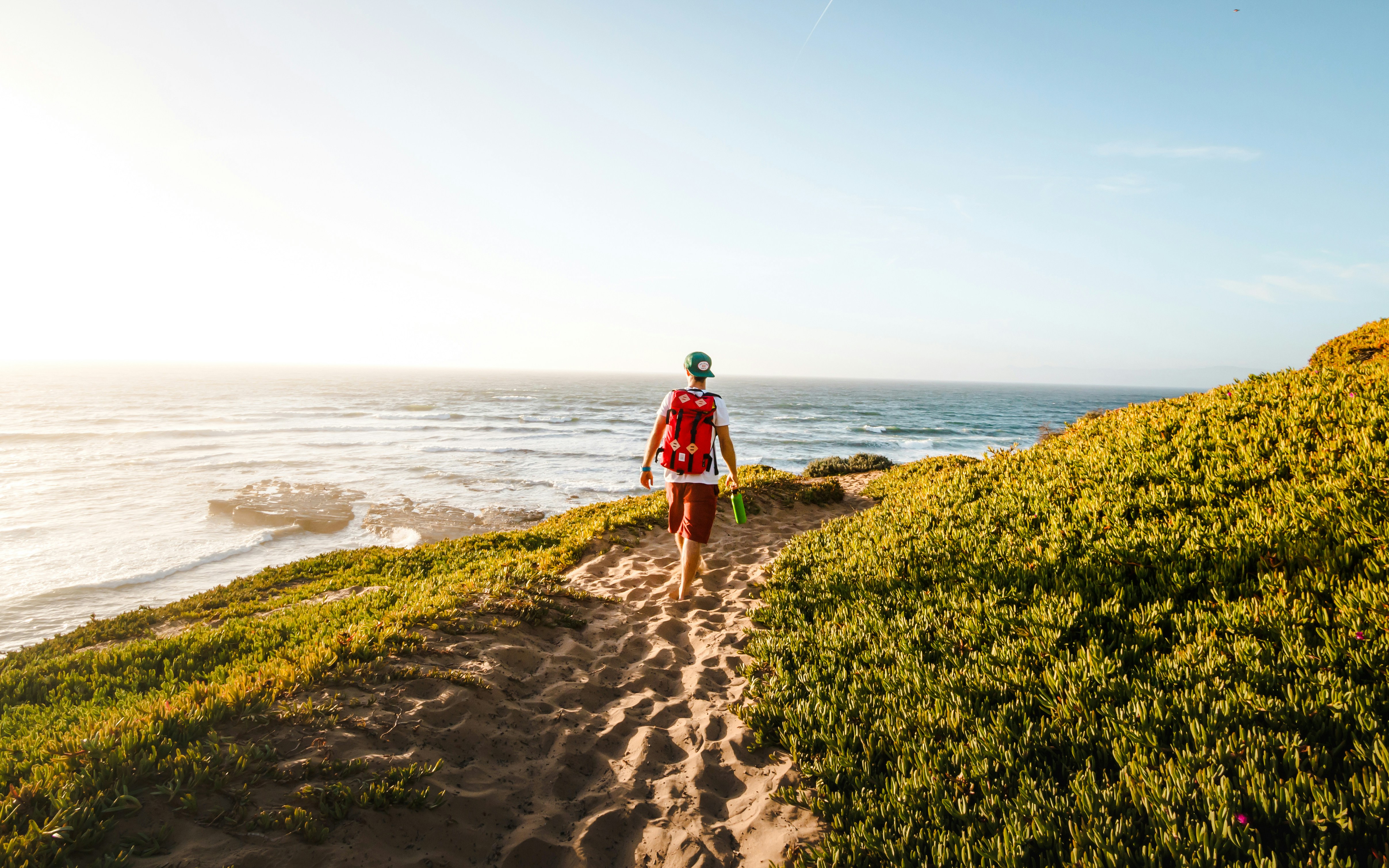 Man hiking along a sandy path near the ocean with greenery on both sides.