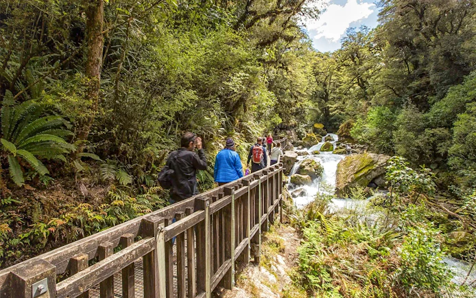 Hikers on Milford Track bridge near waterfall, Te Anau, New Zealand.