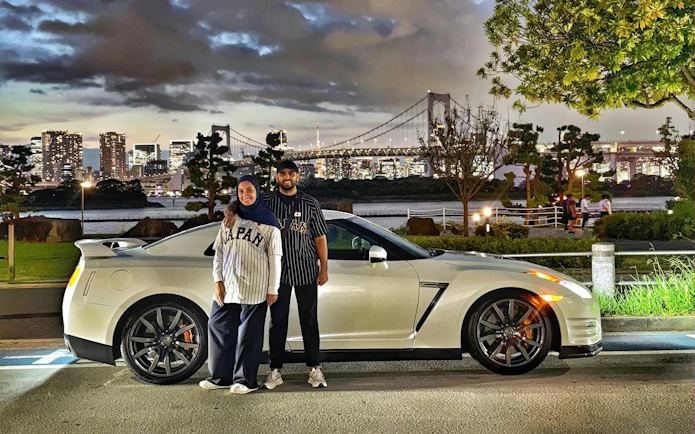 Tokyo skyline with Rainbow Bridge behind a Nissan GT-R R35, featuring two people enjoying a JDM experience.