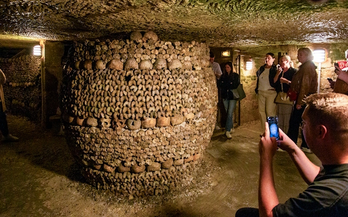 Tourists exploring Paris Catacombs with guide, viewing bone structures in underground passage.