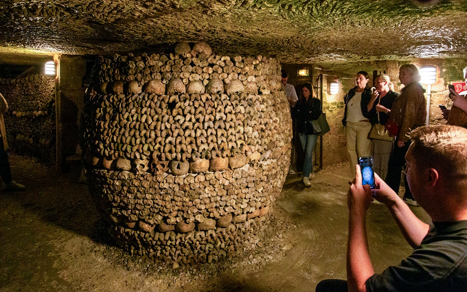 Tourists exploring Paris Catacombs with guide, viewing bone structures in underground passage.