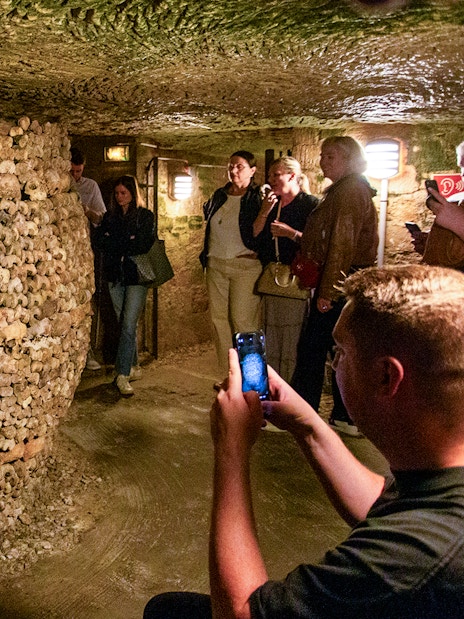 Tourists exploring Paris Catacombs with guide, viewing bone structures in underground passage.