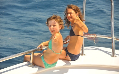 Girls on cruise deck giving thumbs up, watching dolphins in the ocean.