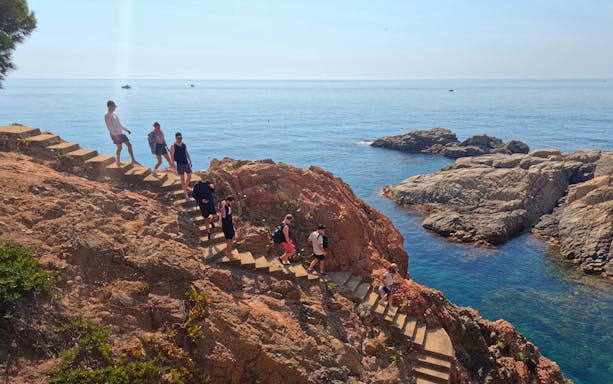 Hikers descending rocky steps on Cami de Ronda coastal path, Costa Brava, Spain.
