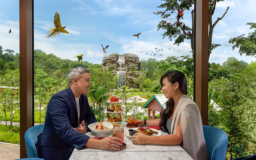 Couple dining at Bird Paradise restaurant with waterfall and birds in view.