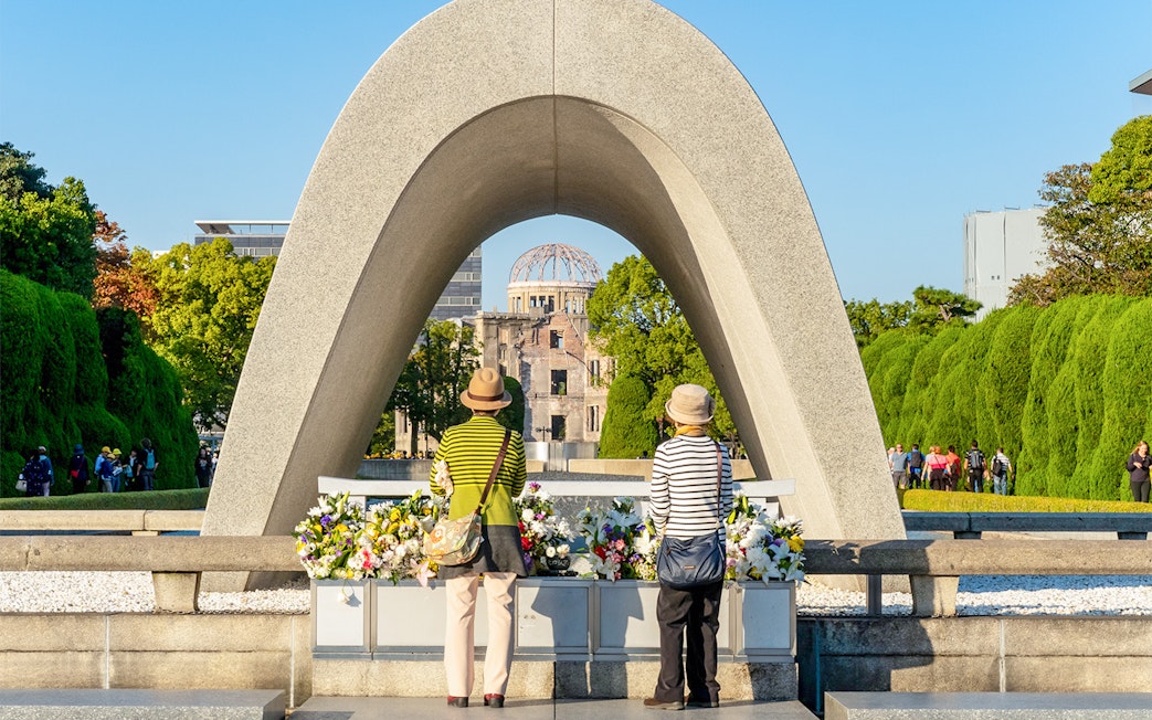 Tourists at Hiroshima Victims Memorial Cenotaph with Atomic Bomb Dome in view.