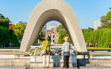 Tourists at Hiroshima Victims Memorial Cenotaph with Atomic Bomb Dome in view.