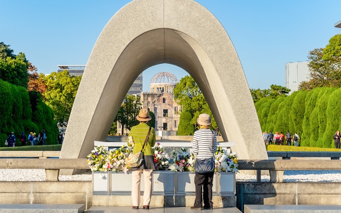 Tourists at Hiroshima Victims Memorial Cenotaph with Atomic Bomb Dome in view.