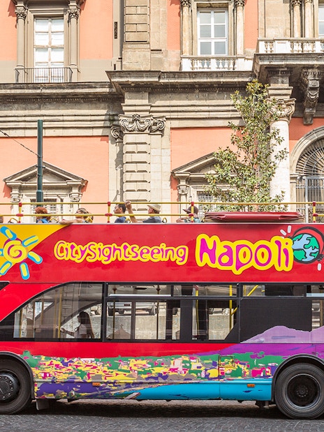 Open-top sightseeing bus in Naples in front of historic building.