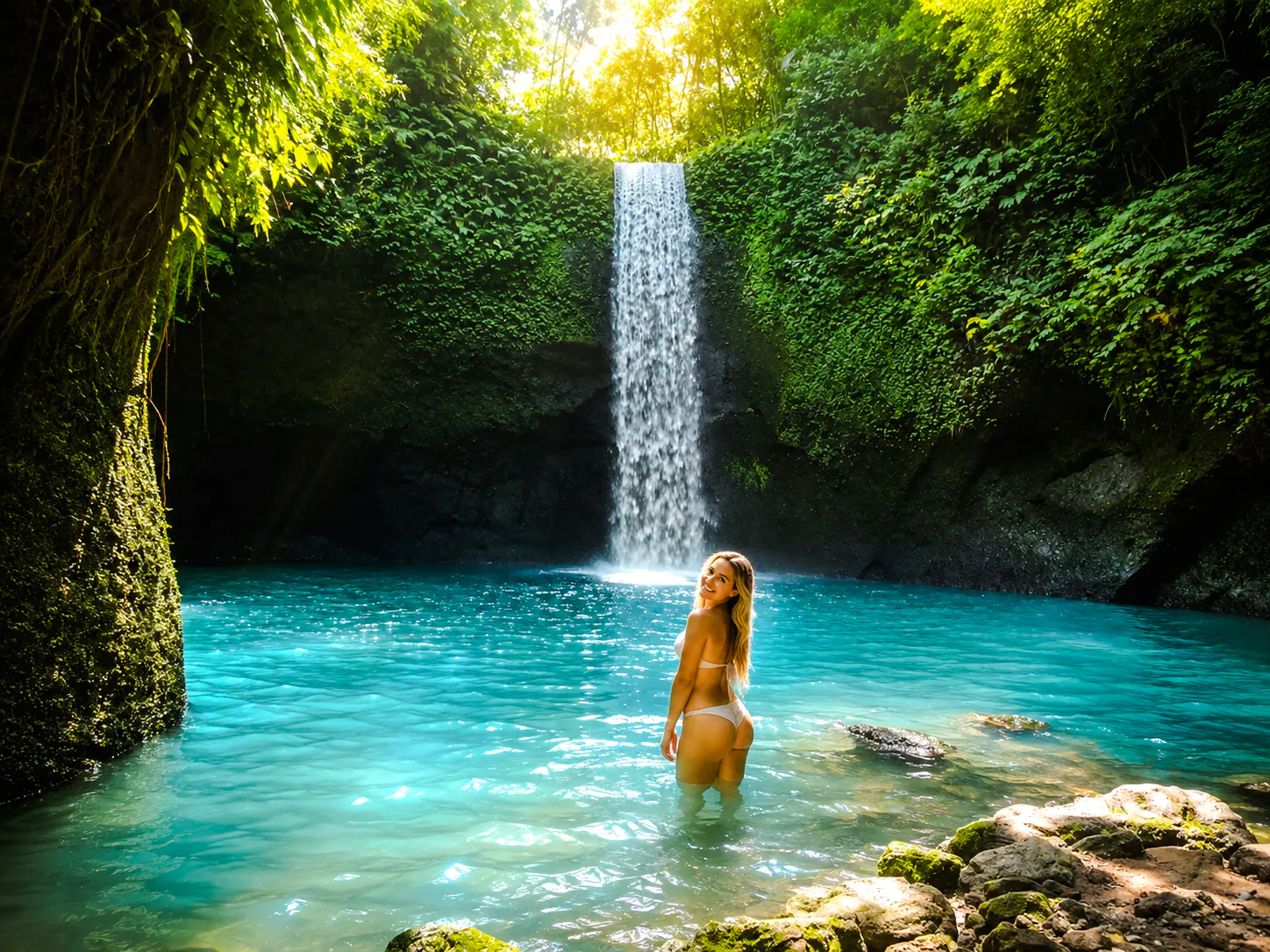Person standing in turquoise pool beneath lush waterfall, Ubud, Bali.