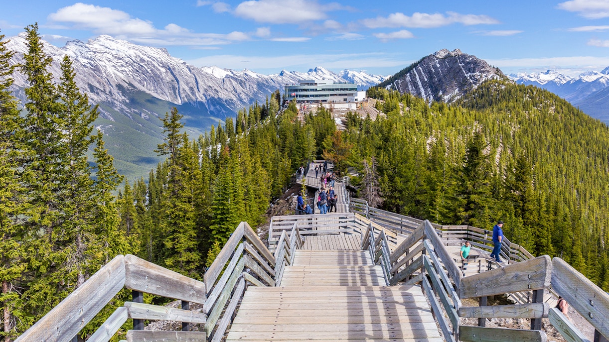Tourists walking on boardwalk at Sulphur Mountain summit, Banff, Canada with mountain views.
