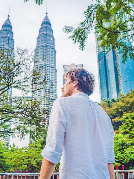 Person standing near Petronas Towers in Kuala Lumpur, surrounded by trees.