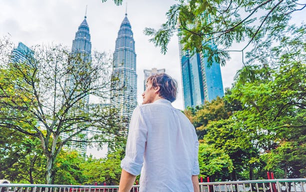 Person standing near Petronas Towers in Kuala Lumpur, surrounded by trees.