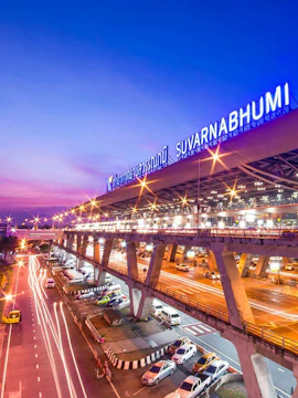 Suvarnabhumi Airport exterior with cars and illuminated terminal at dusk, Bangkok.