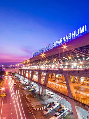 Suvarnabhumi Airport exterior with cars and illuminated terminal at dusk, Bangkok.
