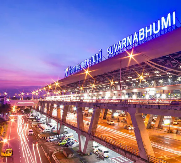 Suvarnabhumi Airport exterior with cars and illuminated terminal at dusk, Bangkok.
