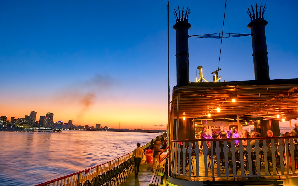 Steamboat Natchez cruising on the Mississippi River at sunset with New Orleans skyline.