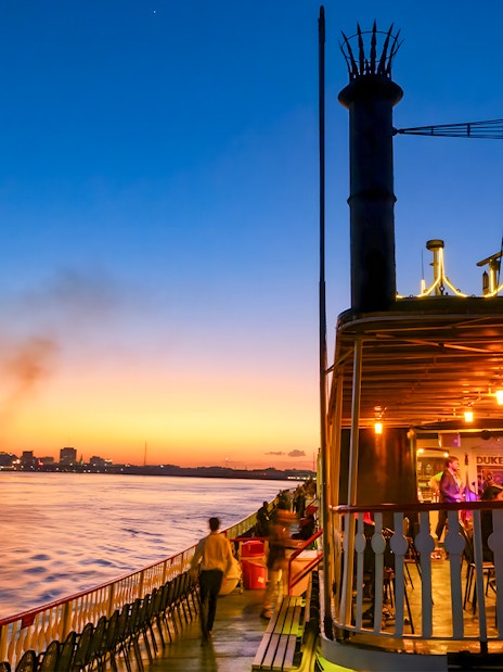 Steamboat Natchez cruising on the Mississippi River at sunset with New Orleans skyline.