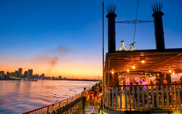 Steamboat Natchez cruising on the Mississippi River at sunset with New Orleans skyline.