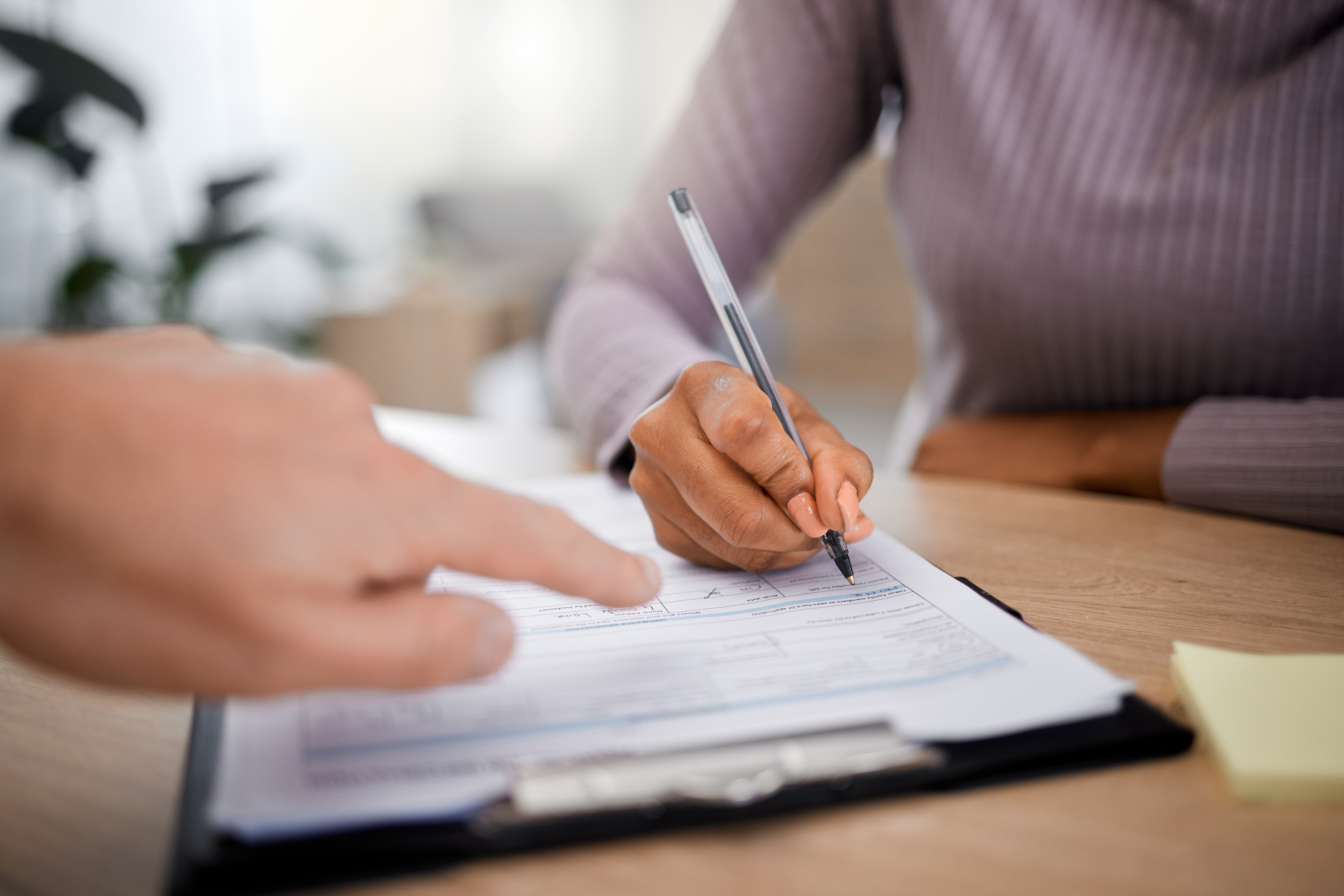 Person signing a document with a pen while another hand points at the paper.