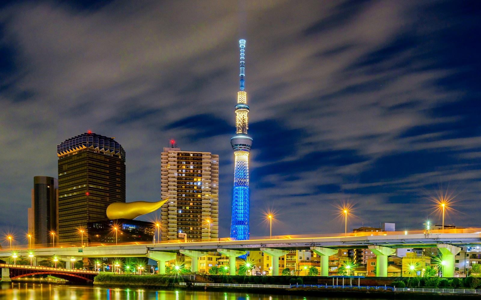 Der beleuchtete Tokyo Skytree bei Nacht, umgeben von modernen Gebäuden und einer Brücke im Vordergrund